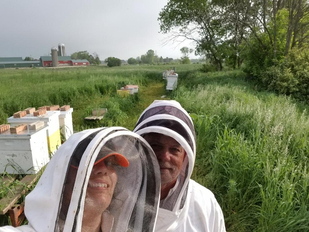 bee+yard Beekeepers wearing protective gear pose for a selfie in a lush green field with beehives in the background, showcasing sustainable agriculture and apiculture practices.
